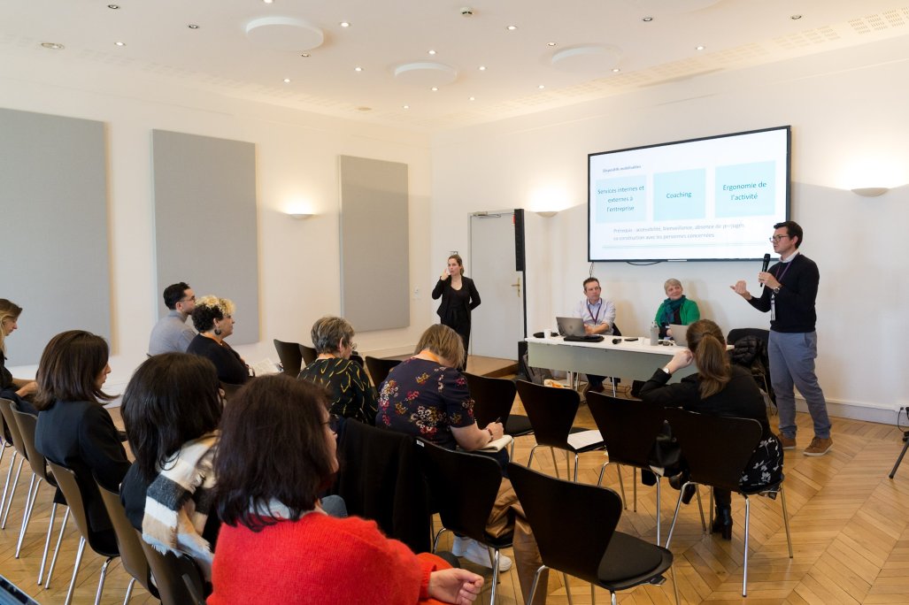 URRH 2023 - Photo des participants de l'atelier LSF. Au premier plan, le public, assis sur des chaises. Dans le fond, une personne debout (à droite) prend la parole avec un micro, 2 personnes au centre, assises, écoutent cette personne et une 4ème, à gauche et debout, traduit en langue des signes française (LSF).