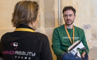 URRH 2023 - Photo de deux participants à l'événement. De dos et assise, une personne qui porte un t-shirt où il est écrit "Handireality". En face, un homme la regarde, il tient, à la main, un casque de réalité virtuelle augmentée.