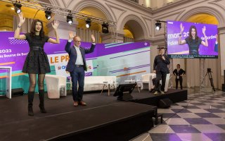 URRH 2023 - Photo de la cérémonie de remise du prix ADP, démonstration de la LSF. De gauche à droite : Sophie Vouzelaud, maitresse de cérémonie en train d'applaudir en Langue des signes françaises, Hugues Defoy, de l'Agefiph, faisant de même, Béatrice, la voix de Sophie pendant la soirée, et au fond à droite l'écran où apparaît Sophie Vouzelaud en grand, pour le public.