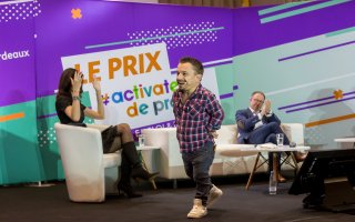 URRH 2023 - Photo de la cérémonie de remise du prix ADP, sketch de Krystoff Fluder. De gauche à droite : Sophie Vouzelaud, assise et en train d'applaudir en LSF, Krystoff Fluder debout en milieu de scène et Hugues Defoy, assis en train d'applaudir.