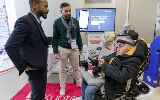 Photo de l'espace exposant. 2 personnes de Kunto parlent à un participant. Sur l'écran en arrière plan est écrit : Des programmes d'entrainement adaptés aux capacités de chacun.