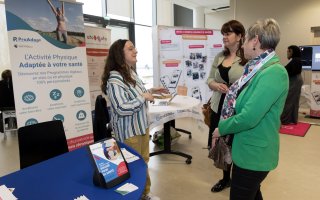 Espace exposant, Pro Adapt. 2 personnes posent des questions à une exposante, au premier plan. En arrière plan apparaissent les stands d'Atomota et de Kunto.