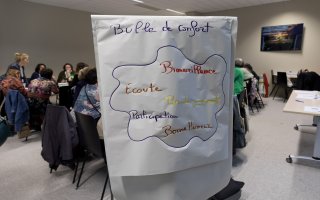 Photo d'un paperboard au premier plan, dans une salle d'atelier remplie de personnes. Il est écrit sur le papier "Bulle de confort : bienveillance, écoute, pas de jugement, participation, bonne humeur.