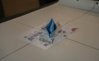 Sur une table, se trouve une feuille avec des dessins (une ancre de bateau, des matelots, une île) et sur cette feuille, un bateau bleu, fait en maquette papier
