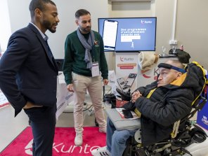 Photo de l'espace exposant. 2 personnes de Kunto parlent à un participant. Sur l'écran en arrière plan est écrit : Des programmes d'entrainement adaptés aux capacités de chacun.