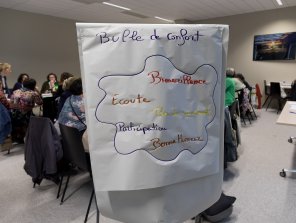 Photo d'un paperboard au premier plan, dans une salle d'atelier remplie de personnes. Il est écrit sur le papier "Bulle de confort : bienveillance, écoute, pas de jugement, participation, bonne humeur.