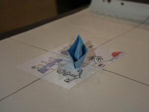 Sur une table, se trouve une feuille avec des dessins (une ancre de bateau, des matelots, une île) et sur cette feuille, un bateau bleu, fait en maquette papier
