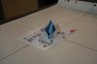 Sur une table, se trouve une feuille avec des dessins (une ancre de bateau, des matelots, une île) et sur cette feuille, un bateau bleu, fait en maquette papier