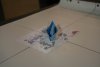 Sur une table, se trouve une feuille avec des dessins (une ancre de bateau, des matelots, une île) et sur cette feuille, un bateau bleu, fait en maquette papier